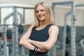 Portrait of adult blonde fitness woman personal trainer with folded hands in gym, beautiful smiling female looking in camera