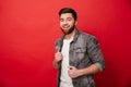 Portrait of affable kind man 30s in jeans jacket posing on camera with amazing smile, isolated over red background Royalty Free Stock Photo