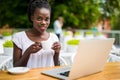 Portrait of African American girl in glasses sitting at the table of cafe with laptop and holding cup of coffee in hands. Royalty Free Stock Photo