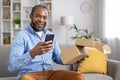 Portrait of an African-American man sitting on the sofa at home, holding an open parcel box in his hand, using a mobile Royalty Free Stock Photo