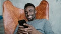 Portrait of African man sitting in chair, using Smartphone. Man reads messages and laughs. Guy typing massages, smiling