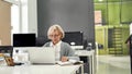 Portrait of aged woman in glasses, senior intern looking focused at the screen while using laptop, sitting at desk Royalty Free Stock Photo