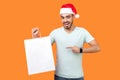 Portrait of amazed bearded young man in santa hat and casual white t-shirt standing pointing at white bag, looking surprised about Royalty Free Stock Photo