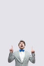 Portrait of amazed handsome bearded man in casual grey suit and blue bow tie standing looking and pointing at up copyspace