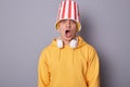 Portrait of amazed shocked emotional hipster man wears yellow hoodie standing against grey wall expressing astonishment, posing
