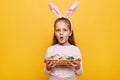 Portrait of amazed surprised little girl wearing bunny ears holding Easter eggs in wicker basket isolated on yellow background, Royalty Free Stock Photo