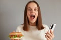 Portrait of amazed young adult caucasian woman wearing white t-shirt holding american classic fast food burger, using mobile phone Royalty Free Stock Photo