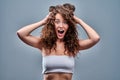 Portrait of amazed young woman with curly hair posing over grey background, keeping palms on her head and looking cheerfully at Royalty Free Stock Photo