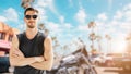 Portrait of American young handsome man chopper biker rider in sleeveless shirt with sunglasses summer coast beach background