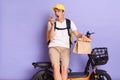 Portrait of angry courier man wearing white t shirt and cap delivering parcels on electric bike isolated over purple background,