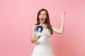 Portrait of angry irritated bride woman in wedding dress screaming in megaphone and spreading hands on pink