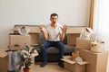 Portrait of angry man wearing white T-shirt and jeans sitting on sofa surrounded with cardboard boxes, raised arms, screaming with