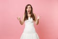 Portrait of angry shocked bride woman with opened mouth in white wedding dress standing spreading hands isolated on pink