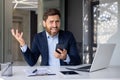 Portrait of an angry young businessman sitting at a desk in the office, holding a phone in his hand and looking Royalty Free Stock Photo