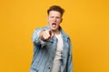 Portrait of angry young man in denim casual clothes pointing index finger on camera, swearing isolated on yellow orange Royalty Free Stock Photo
