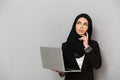 Portrait of arabic woman in black traditional hijab looking upward with thoughtful brooding glance while holding laptop, isolated