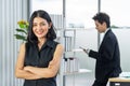 Portrait of asian business woman standing in office with her arms crossed with businessman using phone at the back Royalty Free Stock Photo