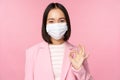 Portrait of asian businesswoman in medical face mask, showing okay sign, wearing suit, work rules during covid-19 Royalty Free Stock Photo