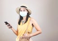 Asian female tourist wearing yellow sleeveless shirt, hat and medical protective mask, holding mobile phone in her hand, smiling Royalty Free Stock Photo