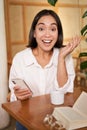 Portrait of asian girl looking surprised, holding mobile phone with amazed face, great news on smartphone, sitting in Royalty Free Stock Photo