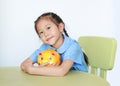 Portrait of Asian little girl in school uniform sitting on table and hugging yellow piggy bank on white background. Schoolgirl Royalty Free Stock Photo