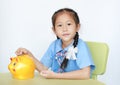 Portrait of Asian little girl in school uniform sitting on table and putting coin into piggy bank isolated on white background. Royalty Free Stock Photo