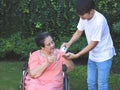 Asian senior woman sitting on wheelchair holding glass of water, get medical pill from her son. garden, outdoor Royalty Free Stock Photo