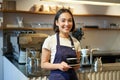 Portrait of asian smiling female barista, cafe waitress holding cup of coffee, serving clients, taking order to the Royalty Free Stock Photo