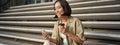 Portrait of asian woman with smartphone, drinks coffee and watches videos on mobile phone. Girl with telephone sits on