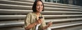 Portrait of asian woman with smartphone, drinks coffee and watches videos on mobile phone. Girl with telephone sits on