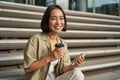 Portrait of asian woman with smartphone, drinks coffee and watches videos on mobile phone. Girl with telephone sits on