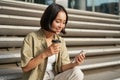 Portrait of asian woman with smartphone, drinks coffee and watches videos on mobile phone. Girl with telephone sits on