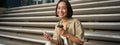 Portrait of asian woman with smartphone, drinks coffee and watches videos on mobile phone. Girl with telephone sits on