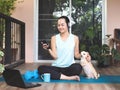 Asian woman wearing headphones, holding coffee cup, using smartphone, sitting  on yoga mat in balcony  with computer laptop Royalty Free Stock Photo