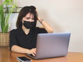 Asian young business woman wearing black protective face mask, sitting at wooden table with computer laptop, touching her head,got Royalty Free Stock Photo