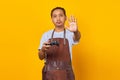 Portrait of Asian young man wearing apron holding game controller and making stop motion with hands over yellow background Royalty Free Stock Photo