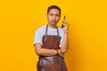 Portrait of Asian young man wearing apron looking angry while talking on smartphone on yellow background