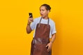 Portrait of Asian young man wearing apron surprised to see incoming message on smartphone on yellow background