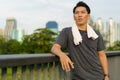 Portrait of Asian young man wearing sportswear, standing and wiping sweat with towel outdoor on walkway with city building Royalty Free Stock Photo