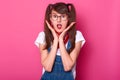 Portrait of astonished emotional girl with pigtails wearing white t shirt, denim overalls and red bandana on neck. Model touches