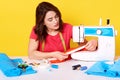 Portrait of attentive good looking tailor sewing clothes, using sewing machine, sitting at table isolated over yellow background