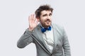 Portrait of attentive handsome bearded man in casual grey suit and blue bow tie standing holding hand near ear and trying to hear