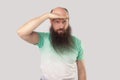 Portrait of attentive middle aged bald man with long beard in light green t-shirt standing with hand on forehead and looking far