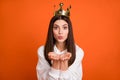 Portrait of attractive amorous girl wearing crown sending air kiss isolated over bright orange color background