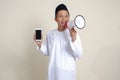 Portrait of attractive Asian muslim man in white shirt showing and presenting blank screen mobile phone while speaking loudly with Royalty Free Stock Photo