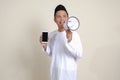 Portrait of attractive Asian muslim man in white shirt showing and presenting blank screen mobile phone while speaking loudly with Royalty Free Stock Photo