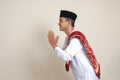 Portrait of attractive Asian muslim man in white shirt with skullcap showing apologize and welcome hand gesture. Isolated image on