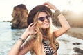 Portrait of attractive blonde girl with long hair posing on rocky beach. She wears bikini, hat, sunglasses. She is Royalty Free Stock Photo