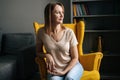 Portrait of attractive blonde woman sitting in yellow chair in dark room with modern interior, thoughtful looking away Royalty Free Stock Photo