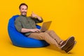 Portrait of attractive cheerful man in bag chair using laptop showing thumbup isolated over bright yellow color Royalty Free Stock Photo